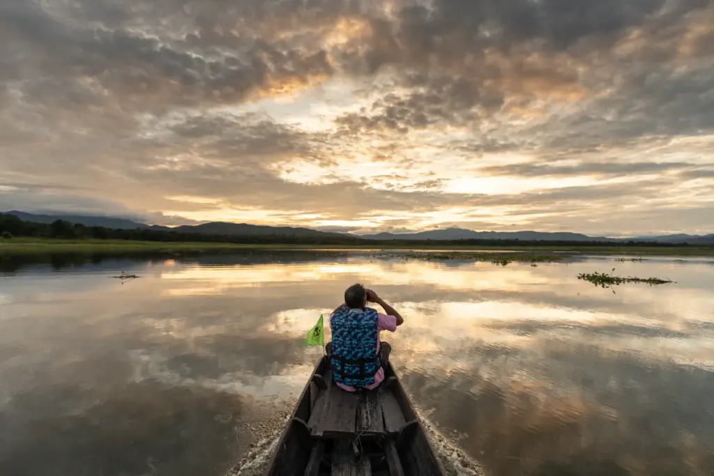 Persoon voorin een houten boot op uitgestrekt water, onder een goudkleurige lucht met wolken.