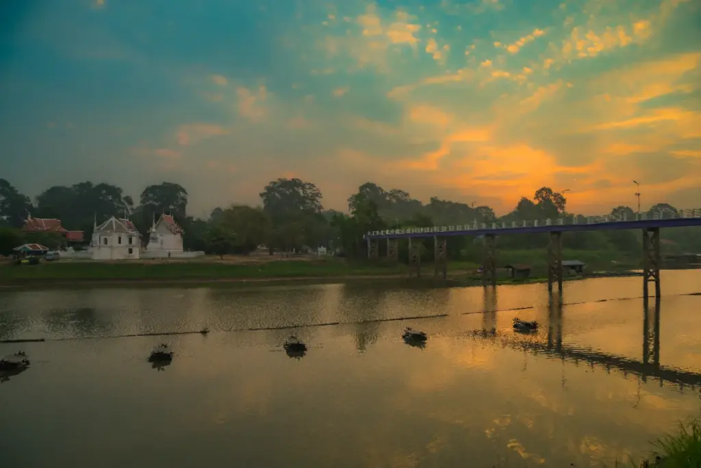 Un pont traverse la rivière Sakae Krang au coucher du soleil, près de bâtiments religieux blancs.