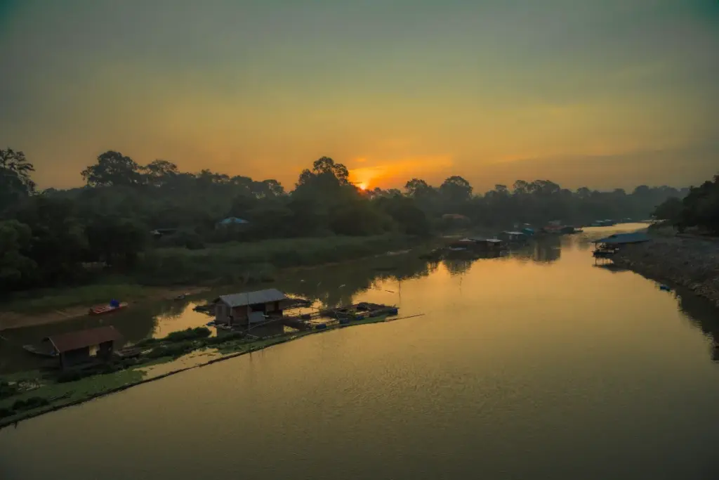 Rivière Sakae Krang au coucher du soleil, avec des maisons flottantes sur l'eau et des arbres sur les berges.