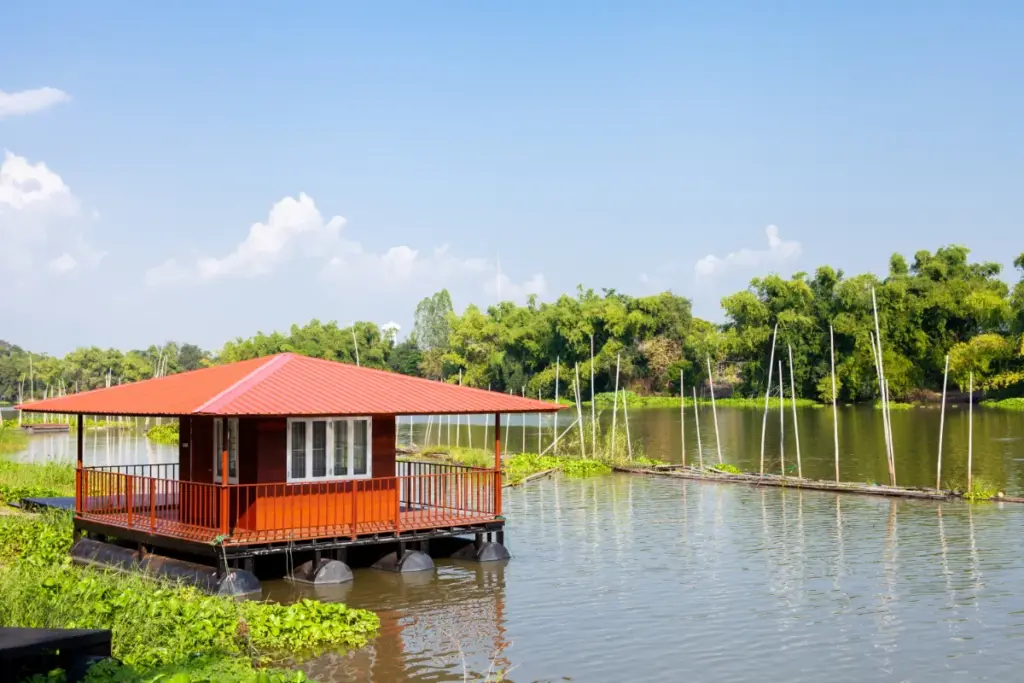Petite maison flottante en bois rouge sur la rivière Sakae Krang, entourée de végétation aquatique.