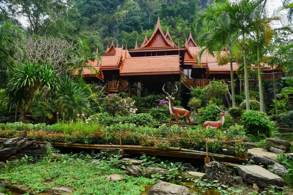 Façade en bois du Wat Tham Khao Wong, entourée de jardins tropicaux fleuris et de sculptures de cerfs.
