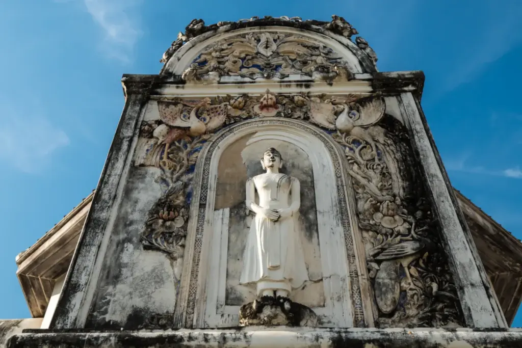 Statue blanche de Bouddha debout dans une niche sculptée de la façade historique du Wat Uposatharam.