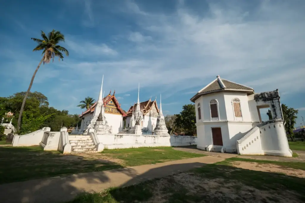 Temple blanc de Wat Uposatharam avec plusieurs chedis, un escalier et un palmier sous le ciel bleu.